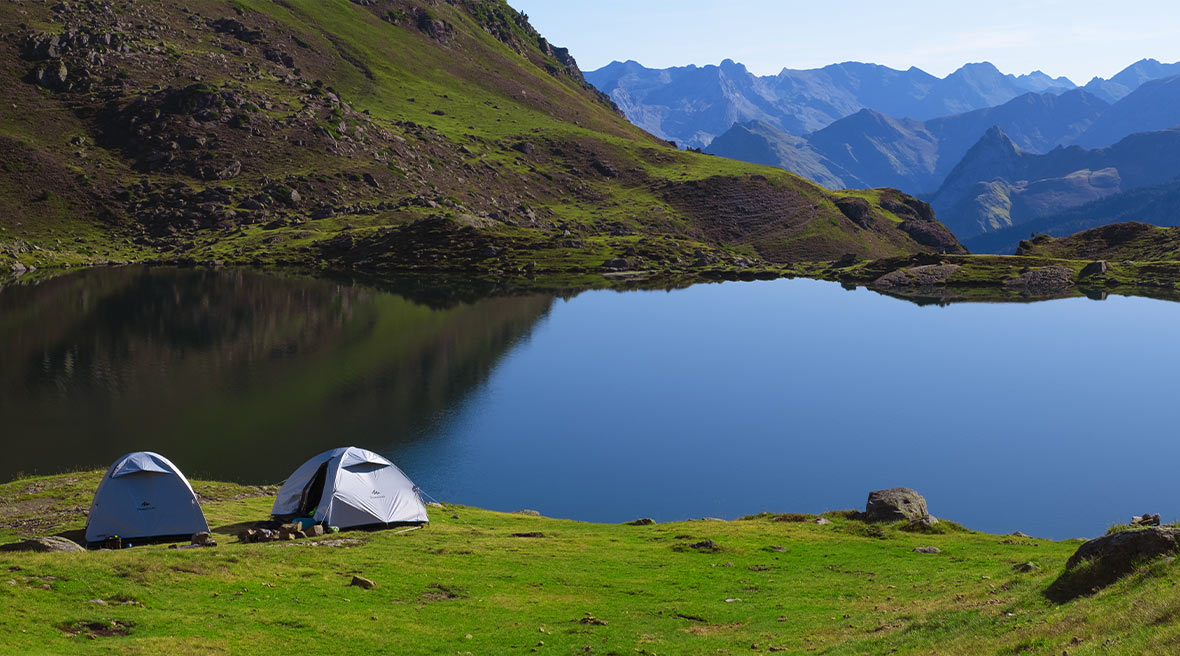 two grey tents pitched by a lake surrounded by the Pyrenees mountains in a lush green park
